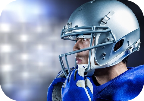 Profile of a focused football player in a blue jersey and silver helmet, gazing ahead. The blurred stadium lights in the background suggest anticipation.